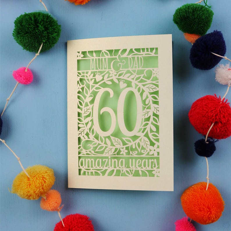 May include: A cream-colored 60th anniversary card with a cut-out floral design. The card reads "Mum & Dad" and "60 amazing years" in a decorative font. The background is light green. Colorful pom-pom garland decorates the blue background.