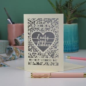 A laser cut "on your wedding day" card with floral design and cut out personalised text. Card features the wedding date, the first names of the couple in a heart shape, and the text "On your wedding day" Standing on a white desk with stationery items in the background. Cut from cream card stock with a silver insert paper