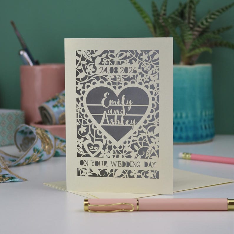A laser cut "on your wedding day" card with floral design and cut out personalised text. Card features the wedding date, the first names of the couple in a heart shape, and the text "On your wedding day" Standing on a white desk with stationery items in the background. Cut from cream card stock with a silver insert paper