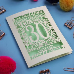 A laser cut keepsake 30th birthday card, personalised with a name in the bottom line of text. Card says Happy Birthday, 30. There is a floral border. Card is cream with a light green insert