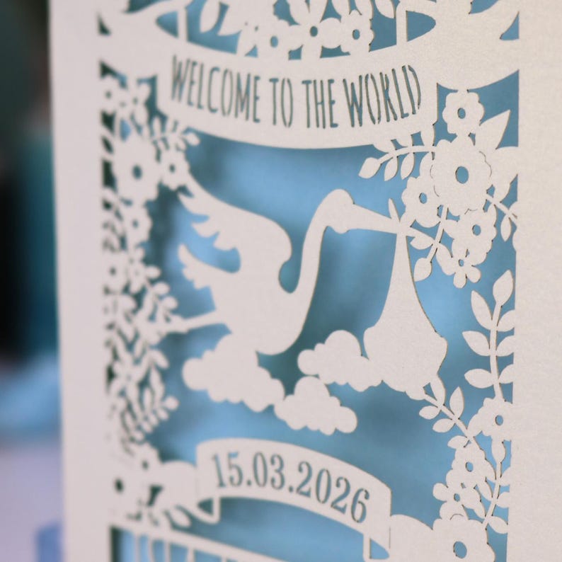 A close up detail of this gorgeous new baby card depicting a stork carrying a bundle surrounded with leaves, flowers and clouds. A banner saying Welcome To The World across the top,  and a second banner below with the baby's date of birth and their name under that. Shown standing on a white desk with stationery items in the fore and background. Its cut from cream cardstock and has a light blue insert paper.