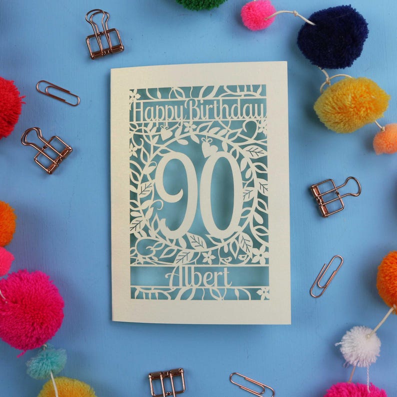 A laser cut keepsake 90th birthday card, personalised with a name in the bottom line of text. Card says Happy Birthday, 90. There is a floral border. Card is cream with a light blue insert
