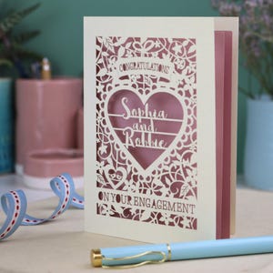 A laser cut engagement card with intricate floral cut out design, the word "Congratulations" in a banner at the top, a heart shape border with 3 lines of text, 'Name and Name', and 'On Your Engagement' in cut out lettering across the bottom of the card.