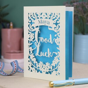 This floral Good Luck card is shown standing on a book with a pen, ribbon and ceramics in the background. It shows the text Good Luck in the centre of the card with a banner above showing the name Maya. It is laser cut from cream card with a peacock blue insert paper
