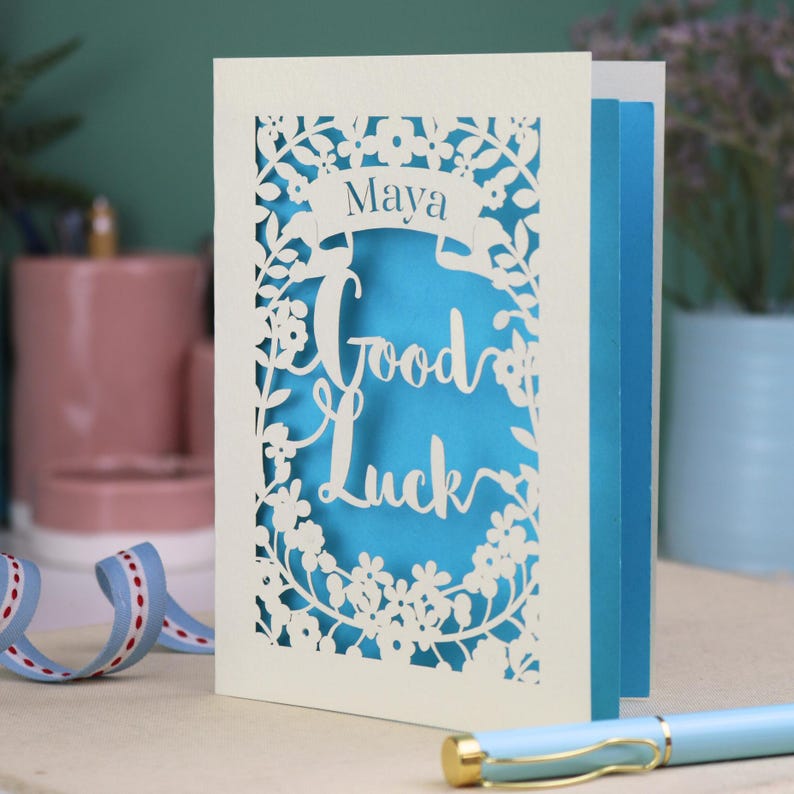 This floral Good Luck card is shown standing on a book with a pen, ribbon and ceramics in the background. It shows the text Good Luck in the centre of the card with a banner above showing the name Maya. It is laser cut from cream card with a peacock blue insert paper