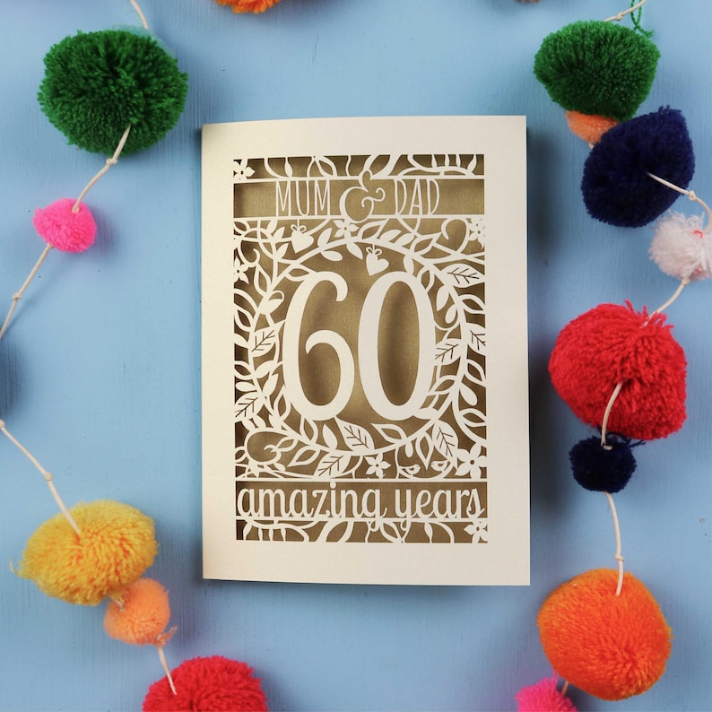 May include: A cream-colored 60th anniversary card with intricate floral cutouts. The card reads "MUM & DAD" at the top and "60 amazing years" at the bottom. Colorful pom-pom garland decorates the blue background.