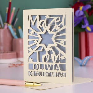May include: A cream-coloured laser-cut greeting card featuring a Star of David, star accents, and the text Mazel Tov! OLIVIA ON YOUR BAT MITZVAH. The card has a blue inner layer visible through the cutouts and stands upright on a flat surface.