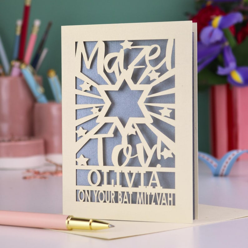 May include: A cream-coloured laser-cut greeting card featuring a Star of David, star accents, and the text Mazel Tov! OLIVIA ON YOUR BAT MITZVAH. The card has a blue inner layer visible through the cutouts and stands upright on a flat surface.