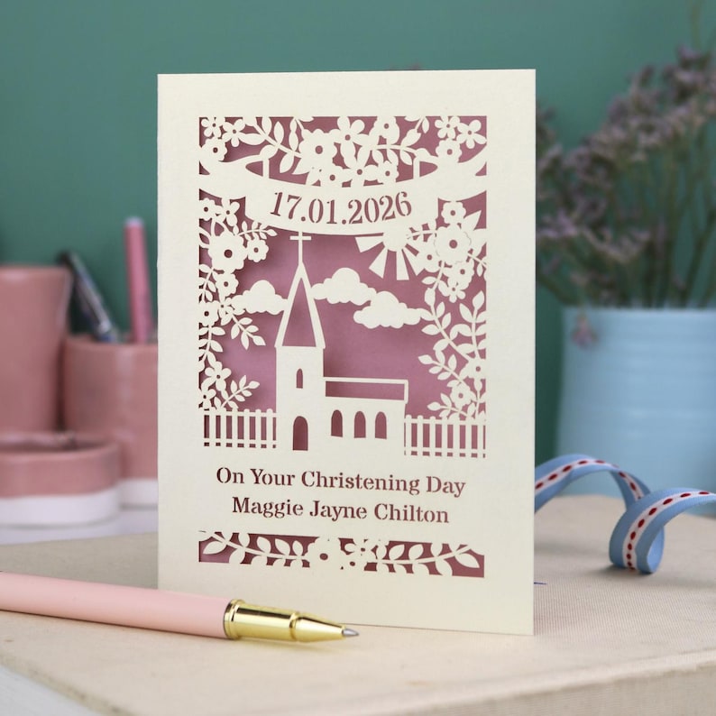 This beautiful laser cut Christening card shows a church surrounded by trees, flowers, clouds and the sun. With a banner flying above the church for the Christening date and beneath the text "On Your Christening Day" and the child's name. Shown standing on a white desk with stationery items, laser cut from cream cardstock with a dusky pink insert paper