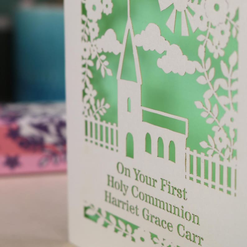 A close up detail of this beautiful laser cut First Holy Communion card shows a church surrounded by trees, flowers, clouds and the sun. With a banner flying above the church for the  Communion date and beneath the text "On Your First Holy Communion" and the child's name. Shown standing on a white desk with stationery items, laser cut from cream cardstock with a light green insert paper