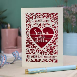 A laser cut engagement card with intricate floral cut out design, the word "Congratulations" in a banner at the top, a heart shape border with 3 lines of text, 'Name and Name', and 'On Your Engagement' in cut out lettering across the bottom of the card.