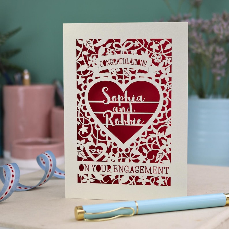 A laser cut engagement card with intricate floral cut out design, the word "Congratulations" in a banner at the top, a heart shape border with 3 lines of text, 'Name and Name', and 'On Your Engagement' in cut out lettering across the bottom of the card.