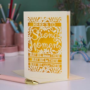 K&ouml;nnte beinhalten: Eine cremefarbene, lasergeschnittene Gru&szlig;karte mit einer leuchtend gelben Einlage. Die Karte zeigt florale Muster und den Text: HERE'S TO Strong Women, MAY WE know THEM, MAY WE be THEM, MAY WE raise THEM. Ein hellrosa Stift liegt im Vordergrund.
