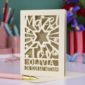 May include: A cream-coloured laser-cut greeting card featuring a Star of David, star shapes, and the text "Mazel Tov! OLIVIA ON YOUR BAT MITZVAH" in a decorative cutout style. The card is positioned upright on a flat surface with a pen in the foreground.