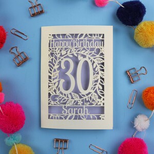 A laser cut keepsake 30th birthday card, personalised with a name in the bottom line of text. Card says Happy Birthday, 30. There is a floral border. Card is cream with a lilac insert