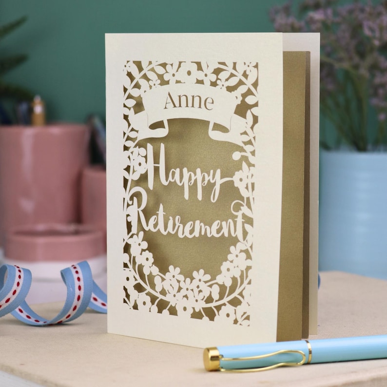 This lovely laser cut retirement card is shown standing on a white desk with a pen and ceramics in the background. It has the words Happy Retirement cut in a script font, has a banner above for the retiree's name and a floral decorative border. It is cut from cream cardstock and has a gold insert paper