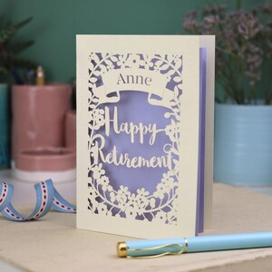 This lovely laser cut retirement card is shown standing on a white desk with a pen and ceramics in the background. It has the words Happy Retirement cut in a script font, has a banner above for the retiree's name and a floral decorative border. It is cut from cream cardstock and has a lilac insert paper