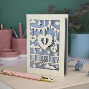 May include: A cream-colored, laser-cut baby announcement card with the name "Theodore Lucas". The card features a heart with footprints, clouds, stars, and banner details. A pink pen with gold accents rests in the foreground.