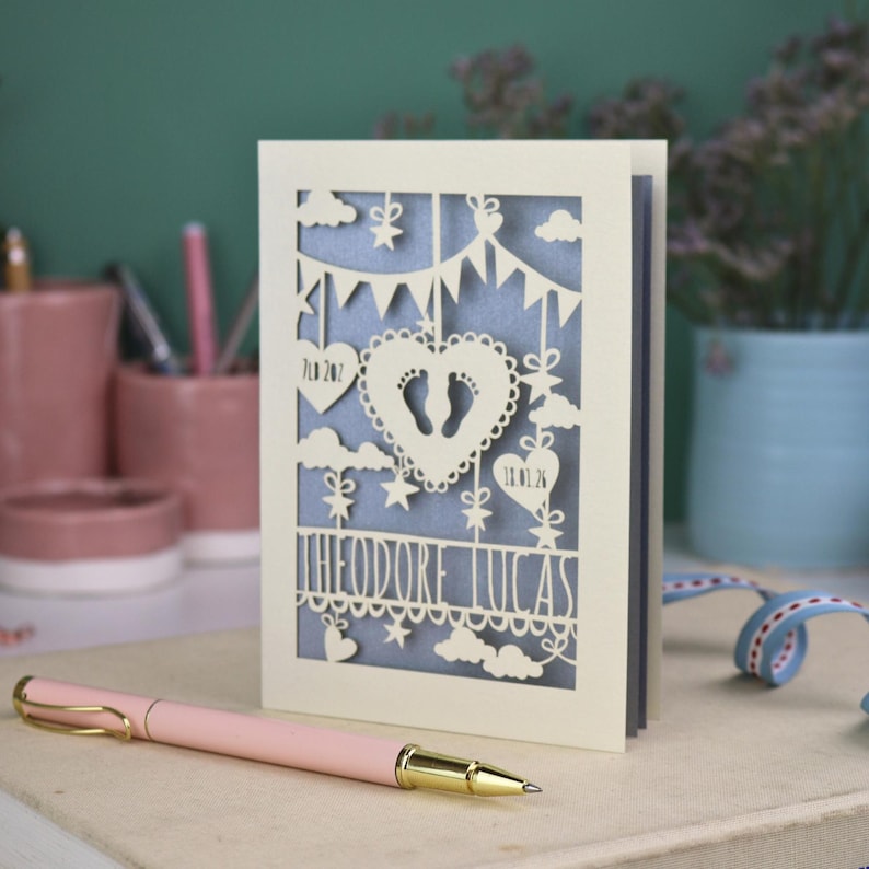 May include: A cream-colored, laser-cut baby announcement card with the name "Theodore Lucas". The card features a heart with footprints, clouds, stars, and banner details. A pink pen with gold accents rests in the foreground.