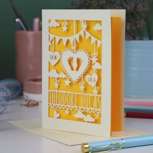 A laser cut new baby card on a white desk with stationery items in the 
background. This cute card is laser cut from a cream cardstock with a sunshine yellow paper insert and features a heart with a pair of baby feet cut out, bunting, clouds, stars and hearts. The text under the heart is the baby's name Theodore Lucas and the left hanging heart shows the baby's weight, 7lb 2oz, and the right hanging heart shows the baby's date of birth 18/01/26