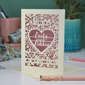 A laser cut "on your wedding day" card with floral design and cut out personalised text. Card features the wedding date, the first names of the couple in a heart shape, and the text "On your wedding day" Standing on a white desk with stationery items in the background. Cut from cream card stock with a dusky pink insert paper