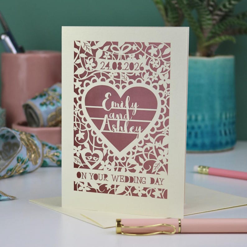 A laser cut "on your wedding day" card with floral design and cut out personalised text. Card features the wedding date, the first names of the couple in a heart shape, and the text "On your wedding day" Standing on a white desk with stationery items in the background. Cut from cream card stock with a dusky pink insert paper
