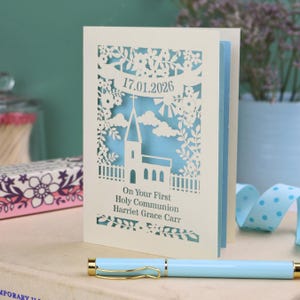 This beautiful laser cut First Holy Communion card shows a church surrounded by trees, flowers, clouds and the sun. With a banner flying above the church for the  Communion date and beneath the text "On Your First Holy Communion" and the child's name. Shown standing on a white desk with stationery items, laser cut from cream cardstock with a light blue insert paper