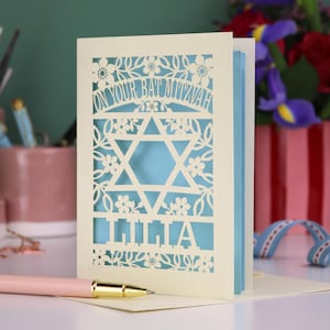 A laser cut Bat Mitzvah card with Star of David design and text "On Your Bat Mitzvah" at the top. The card is personalised with the recipient's name at the bottom and had floral and leaf design elements around the border.