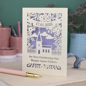 This beautiful laser cut Christening card shows a church surrounded by trees, flowers, clouds and the sun. With a banner flying above the church for the Christening date and beneath the text "On Your Christening Day" and the child's name. Shown standing on a white desk with stationery items, laser cut from cream cardstock with a lilac insert paper
