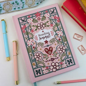 May include: A pink hardback reading journal featuring a dark green floral and bird pattern with a central white label reading Reading Journal. The journal is surrounded by a light blue pen, a pink pen, a pink pencil, and two copper envelope clips.