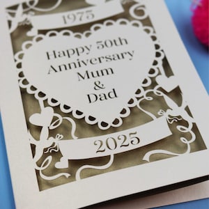 May include: A cream-colored, laser-cut anniversary card. The card features the text "Happy 50th Anniversary Mum & Dad" inside a heart-shaped design. The years 1975 and 2025 are also displayed. Decorative cut-out elements include ribbons and hearts.