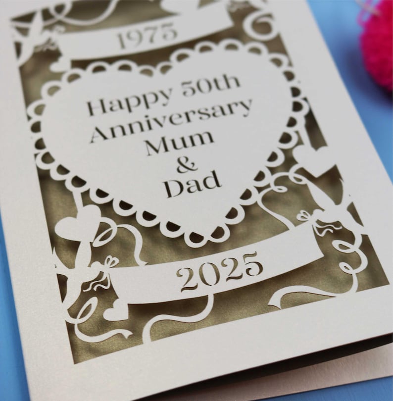 May include: A cream-colored, laser-cut anniversary card. The card features the text "Happy 50th Anniversary Mum & Dad" inside a heart-shaped design. The years 1975 and 2025 are also displayed. Decorative cut-out elements include ribbons and hearts.
