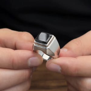 Peut inclure: Une bague en argent avec une grosse pierre d'onyx noire carrée. La bague a un design géométrique avec un large anneau et un sertissage surélevé. La bague est tenue entre deux mains.