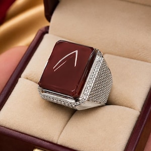 Op de afbeelding: Een zilveren ring met een grote, rechthoekige, donkerrode steen met een wit gegraveerd symbool op het oppervlak. De band is breed, getextureerd met een rasterpatroon en geaccentueerd met kleine, heldere steentjes langs de randen.