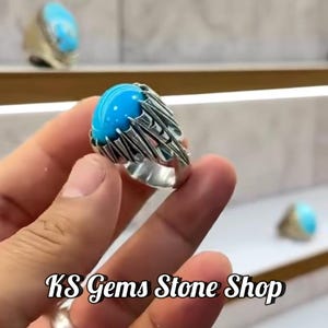 Op de afbeelding: Een zilveren ring met een grote, ovale, helderblauwe steen. De ring heeft een gedetailleerd, gestructureerd ontwerp rond de steen. De tekst "KS Gems Stone Shop" staat onderaan de afbeelding. Andere ringen zijn zichtbaar op de achtergrond.