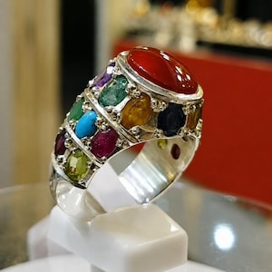 May include: A silver ring featuring a large, oval, polished red stone at the top, surrounded by multiple smaller, multi-coloured oval gemstones including green, blue, yellow, purple, and dark red stones set in a metallic band with a wide profile.