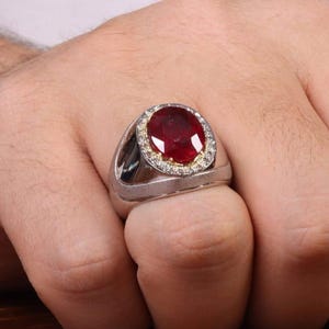 May include: A silver ring featuring a large, oval-cut ruby gemstone. The ruby is surrounded by a halo of smaller, clear gemstones. The ring is set on a silver band, showcasing a classic and elegant design.