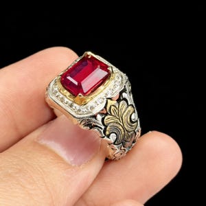 May include: A ring featuring a rectangular, deep red gemstone set in a gold-toned bezel, surrounded by a halo of small clear stones. The band is crafted from silver-toned metal with intricate, raised gold-toned floral or scrollwork detailing.