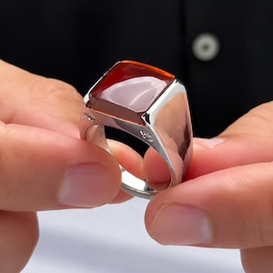 Puede incluir: Un anillo de tono plateado con una gran piedra rectangular pulida de color rojo anaranjado intenso engastada en una banda de metal liso. El anillo tiene un cuerpo ancho y sólido con pequeños grabados decorativos en los laterales.
