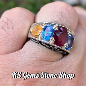 Puede incluir: Un anillo de plata con cuatro piedras preciosas de corte ovalado en naranja, azul brillante, rojo oscuro y azul oscuro, acentuadas con pequeñas piedras claras. La banda tiene detalles negros grabados. El texto KS Gems Stone Shop es visible.