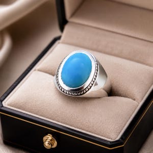 Könnte beinhalten: Ein Silberring mit einem großen, glatten, ovalen Türkisstein in der Mitte. Die Fassung um den blauen Stein herum hat eine detaillierte Perlenstruktur, und der Ring besteht aus poliertem, massivem Silbermetall mit einem breiten, zulaufenden Design.