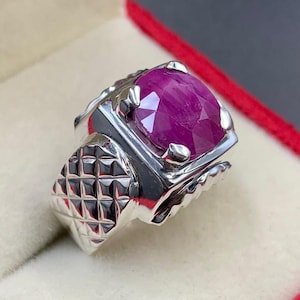 May include: A silver ring featuring a large, faceted, oval-cut ruby gemstone. The ring has a detailed, geometric pattern on the band and a polished finish. The ruby is a deep, vibrant red color.