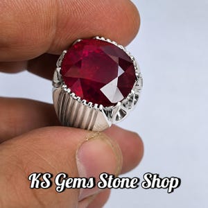 Pode incluir: Um anel de prata com uma grande pedra preciosa redonda, facetada e de cor vermelho-escura, montada numa banda de metal decorativa com ranhuras verticais e detalhes recortados. O texto KS Gems Stone Shop está impresso a preto na imagem.