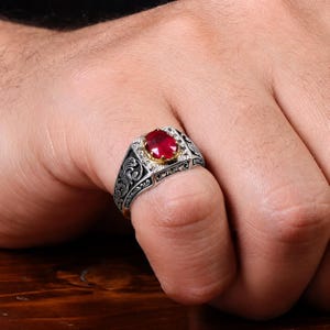 Könnte beinhalten: Ein Silberring mit einem oval geschliffenen roten Edelstein in einer goldfarbenen Fassung. Die Ringschiene ist mit detaillierten, schwarz oxidierten Rankengravuren verziert, die sich entlang der Seiten erstrecken und einen Kontrast bilden.