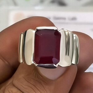 May include: A polished silver-toned ring featuring a large, rectangular, deep red gemstone set in the centre. The band has a wide, smooth, and reflective metallic surface that curves around the finger, framing the dark red stone securely.