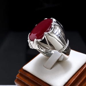 May include: A polished silver ring featuring a large, smooth, oval-shaped deep red gemstone set in a pronged mounting. The band has a reflective metallic finish with intricate structural detailing along the sides and a beaded texture accent.