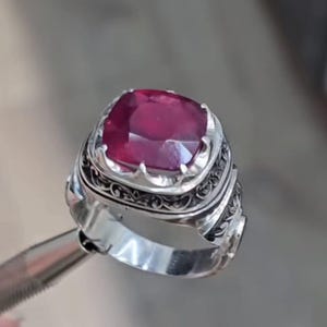 May include: A silver ring with an ornate design, featuring a large, cushion-cut ruby gemstone. The ruby is a deep red color and is set in a detailed, engraved setting. The ring's band has intricate patterns.