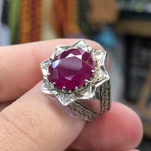 May include: A silver ring featuring a large, oval-cut, deep red ruby gemstone set in a star-shaped frame. The setting is accented with small, clear stones and the band displays intricate, engraved floral patterns along the sides.