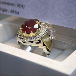 May include: A silver and gold ring featuring a large, oval-cut, deep red gemstone. The gemstone is set in a gold crown-like setting, surrounded by smaller stones. The ring has an ornate design.