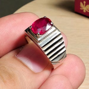 Puede incluir: Un anillo de plata con una piedra preciosa de color rojo intenso de corte ovalado, engastada en cuatro garras. La banda presenta una serie de ranuras horizontales paralelas que se extienden por la superficie superior, creando una textura acanalada.